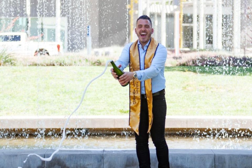 Gennaro Tecchia opening a bottle of champagne while wearing a graduation stole